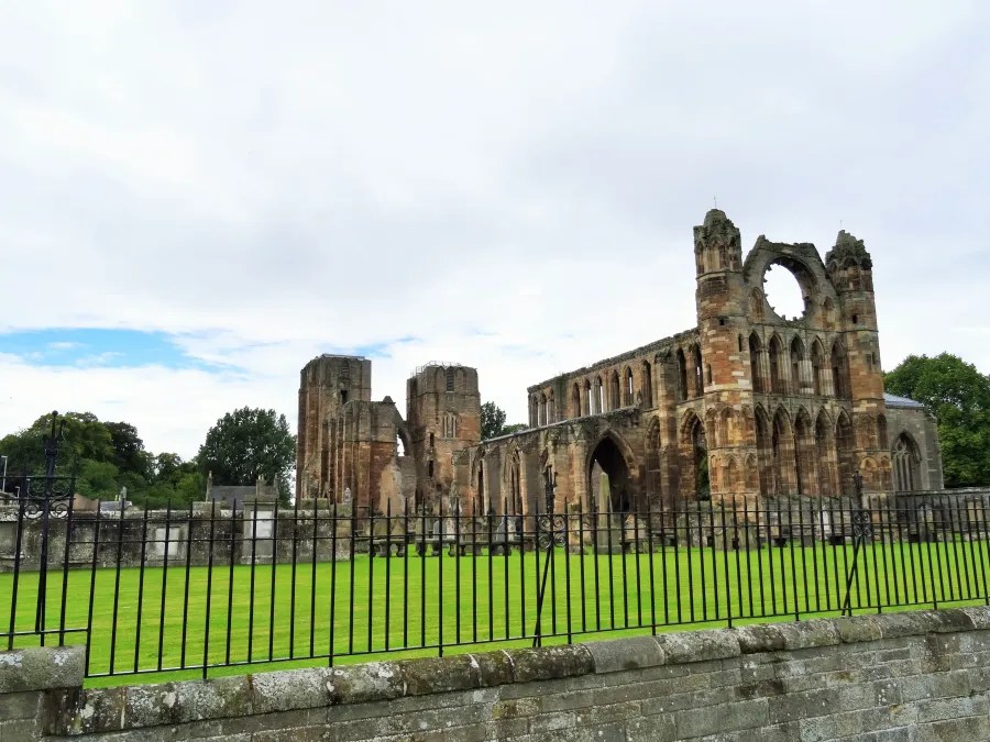 Ruine einer großen Kathedrale mit einem gepflegten grünen Rasen im Vordergrund, umgeben von einem Eisenzaun.