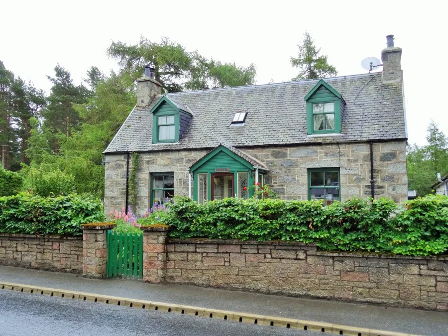 Ein charmantes, zweigeschossiges Steinhaus mit grünem Giebel und Fensterläden, umgeben von dichter Vegetation und einem gewachsenen Gartenzaun.
