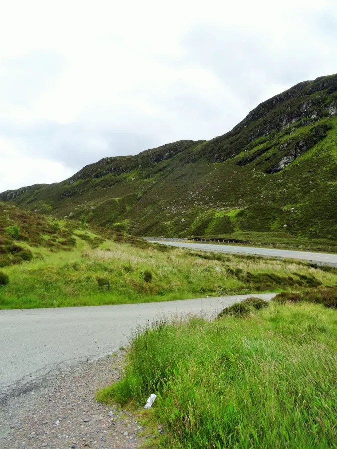 Kurvenreiche Straße durch grüne Highlands mit sanften Hügeln und Wolken am Himmel.