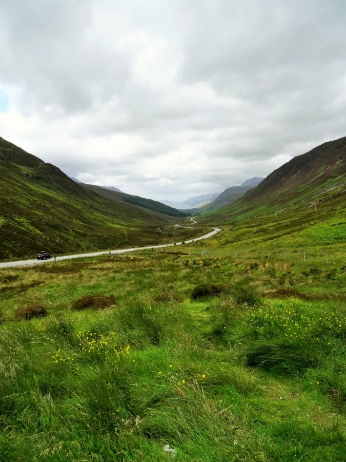 Blick auf eine kurvige Straße, die durch grüne Hügel und Täler in den schottischen Highlands führt, unter einem bewölkten Himmel.