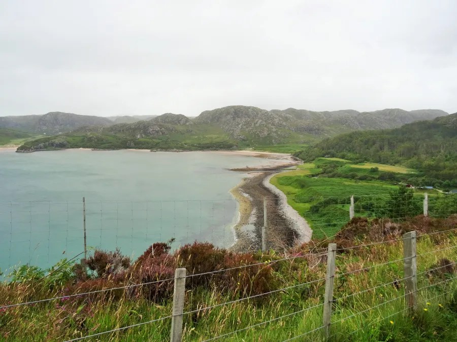 Eine Küstenlandschaft in Schottland mit sanften Hügeln, rauem Wasser und einem bewölkten Himmel, umgeben von grünem Gras und gegen eine Drahtzaun blickend.