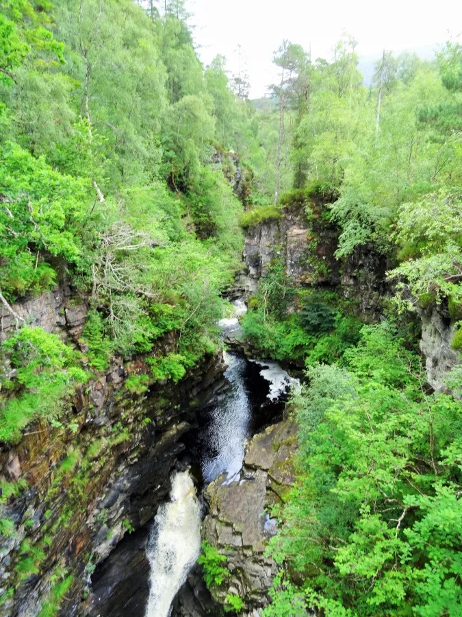 Blick in die Corrieshalloch Gorge, umgeben von üppigem Grün und einem langsam fließenden Wasserfall.
