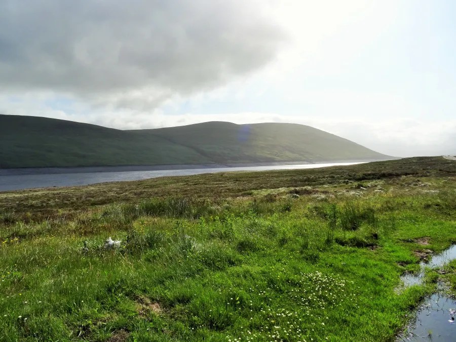 Weite Landschaft mit sanften Hügeln und einem stillen Gewässer im Vordergrund, unter einem bewölkten Himmel.