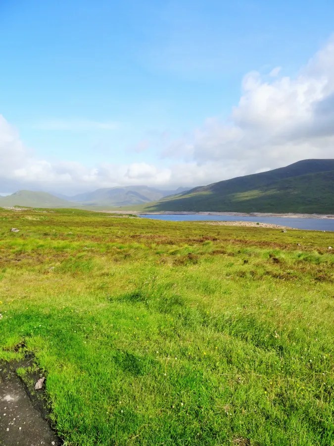 Eine weitläufige Landschaft in Schottland, geprägt von saftigem grünen Gras, sanften Hügeln und einem klaren blauen Himmel. Im Hintergrund ist ein ruhiger See mit umliegenden Bergen zu sehen.