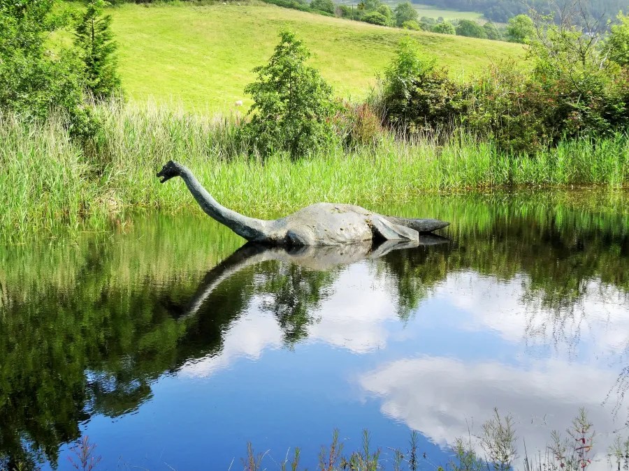 Ein Modell des Loch Ness Monsters, das in einem ruhigen Gewässer liegt, umgeben von grünen Wiesen und Bäumen.