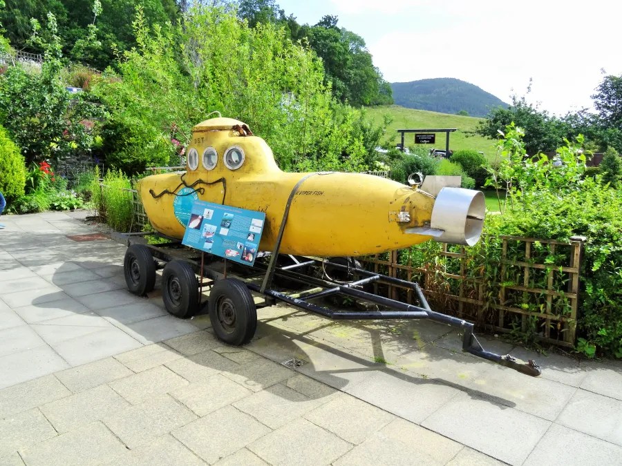 Ein gelbes, U-förmiges Boot steht auf einem Verkehrsanhänger, umgeben von grünen Pflanzen und Bäumen, mit einem Schild vorne, das Informationen bereitstellt.