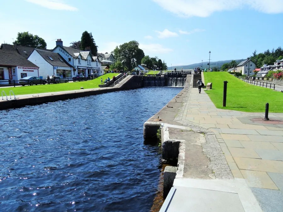 Blick auf den Kanal und die Schleuse in Fort Augustus, Schottland, mit charakteristischen Gebäuden und einem sonnigen Himmel im Hintergrund.