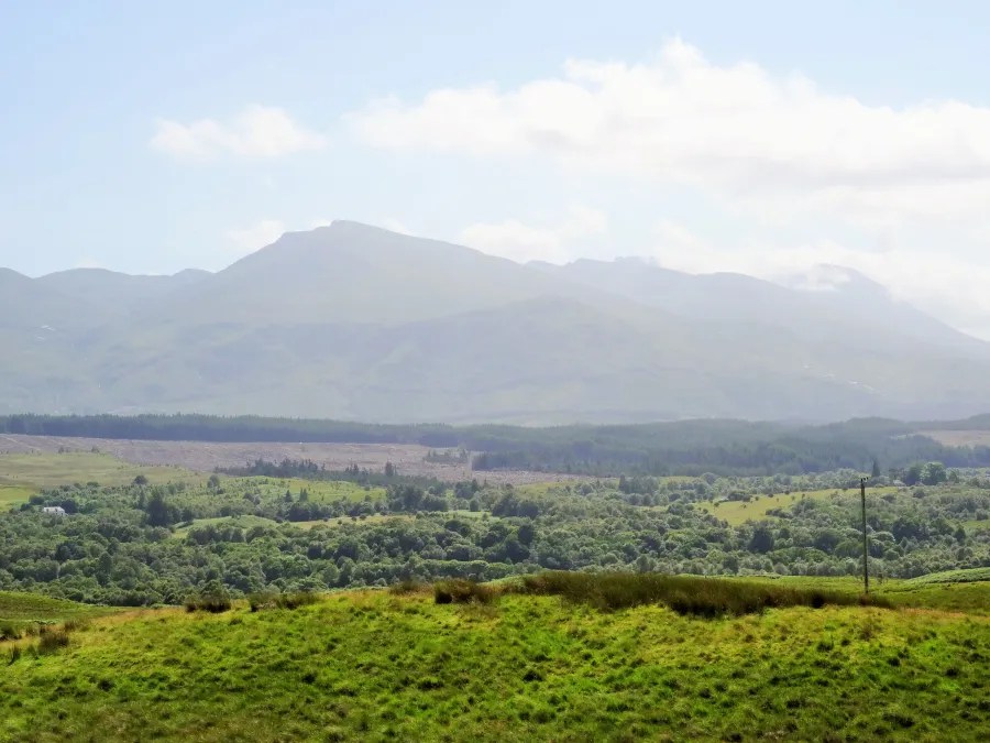 Eine weite Aussicht auf hügelige Landschaften der schottischen Highlands mit grünen Wiesen und Bergen im Hintergrund unter einem hellblauen Himmel.