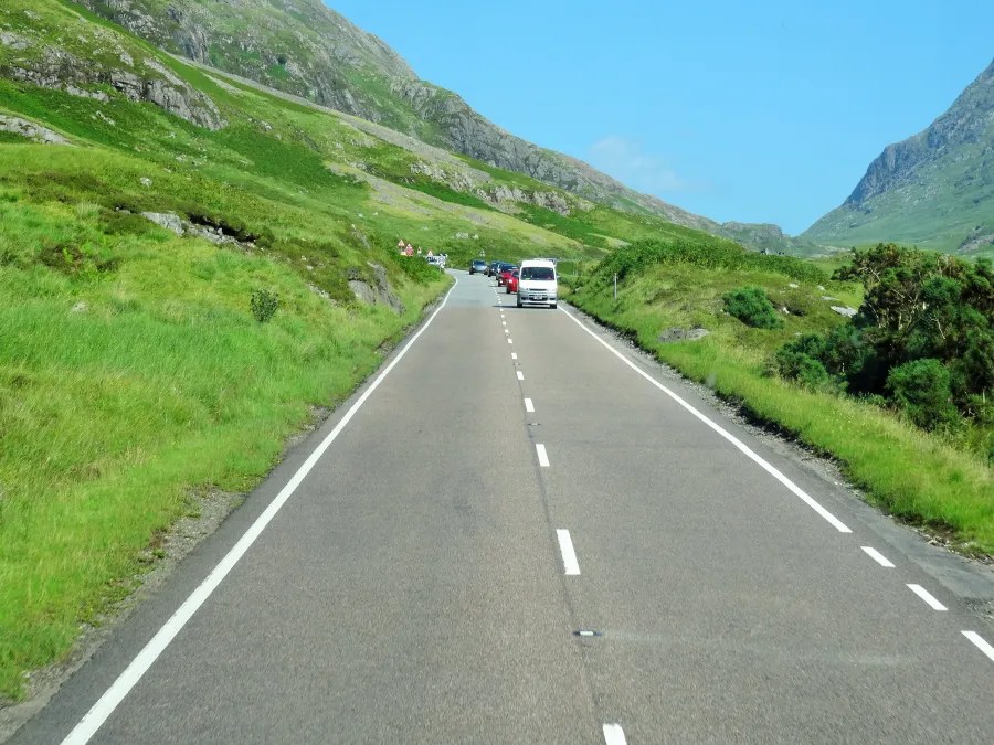 Eine kurvenreiche Straße führt durch die grüne schottische Landschaft, umgeben von Bergen und üppigem Bewuchs.
