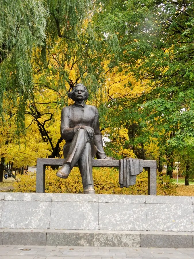 Eine Bronze-Statue von Karl Marx sitzt auf einer Bank, umgeben von buntem Laub im Herbst.