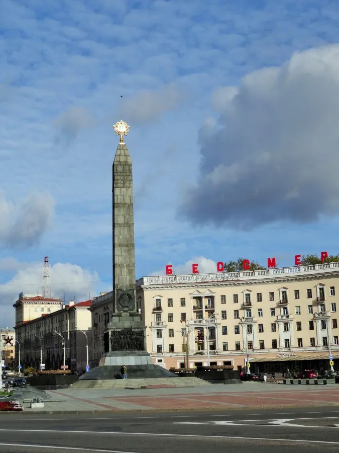 Ein Monument mit goldener Spitze steht auf einem großen Platz in Minsk, umgeben von historischen Gebäuden und unter einem klaren blauen Himmel.