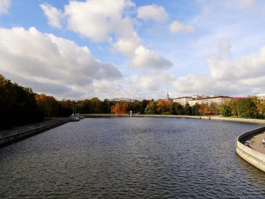 Ein ruhiger Park mit einem großen Gewässer, umgeben von buntem Herbstlaub und Wolken am Himmel.