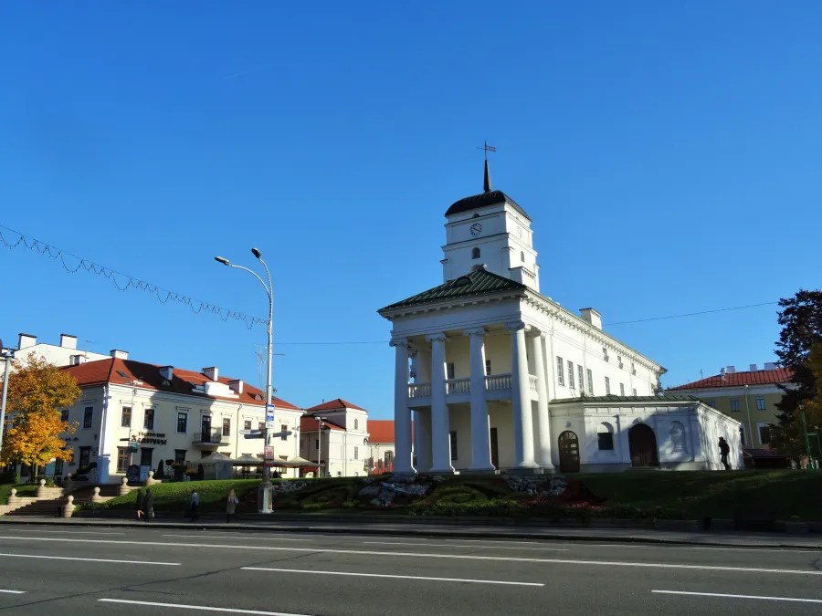 Ein historisches Gebäude mit Säulen und Uhrenturm vor klarem blauen Himmel. Im Vordergrund eine Straße mit wenigen Passanten, während auf der linken Seite benachbarte, mehrstöckige Gebäude im Hintergrund sichtbar sind.