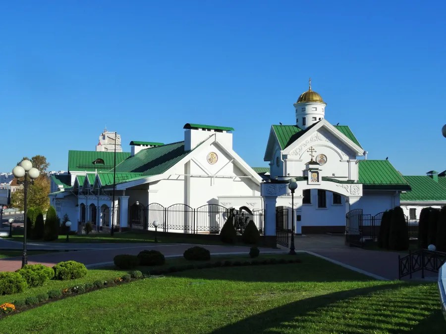 Blick auf ein weißes Gebäude mit grünem Dach und goldener Kuppel, umgeben von Gras und Bäumen, unter blauem Himmel.