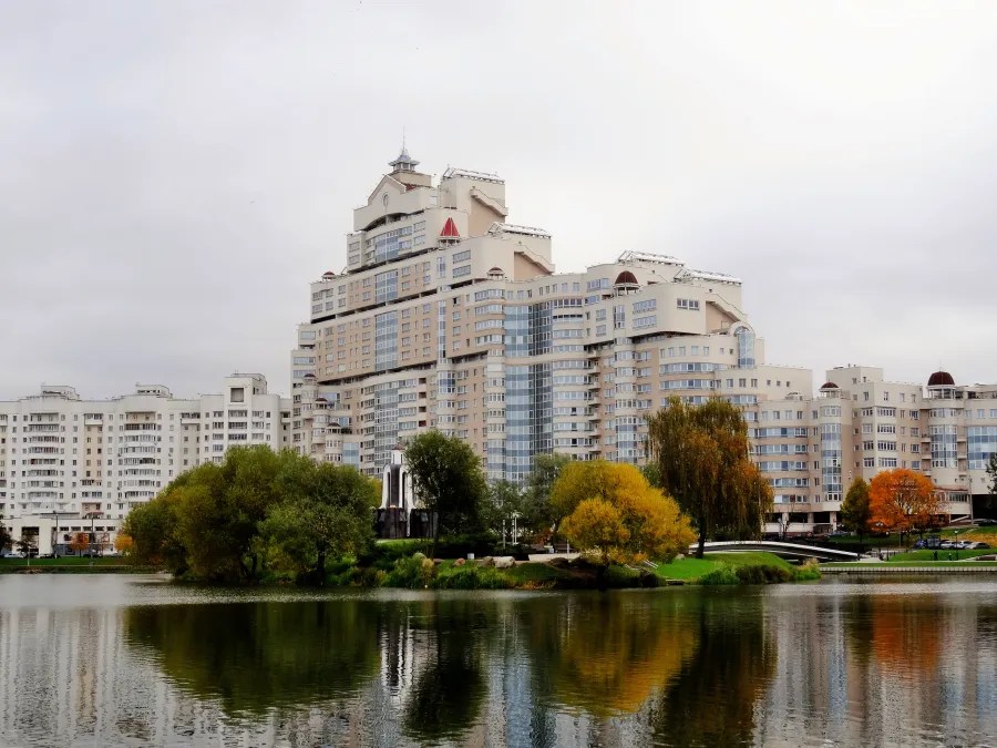Ein modernes Gebäude am Ufer eines ruhigen Gewässers mit Bäumen im Vordergrund und einem bewölkten Himmel.