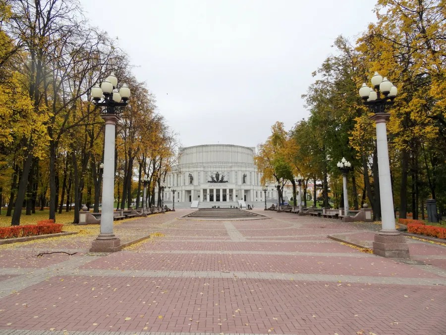 Eine Ansicht des Opernhauses in Minsk, umgeben von herbstlichen Bäumen mit gelben Blättern und Laternen im Vordergrund.