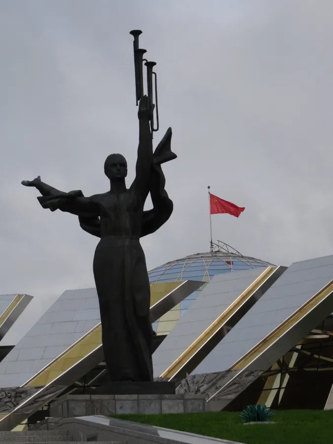 Eine monumentale Statue einer Frau mit einer Trompete, die eine Hand erhebt, vor einem modernen Gebäude mit einem Glaskuppeldach und einer roten Flagge im Hintergrund.