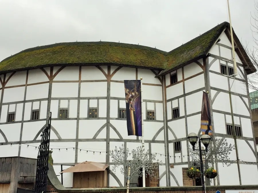 Die Außenseite des Globe Theatre in London, ein historisches Theater, das mit einem grünen Dach und Holzrahmen gestaltet ist. Banner hängen an den Wänden, und es sind dekorative Elemente im Vordergrund zu sehen.