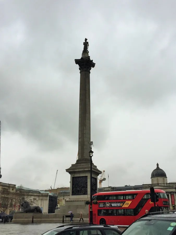 Ein roter Doppeldeckerbus fährt vorbei an der Nelsonsäule auf dem Trafalgar Square. Der Himmel ist grau und es regnet.
