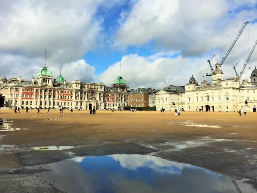 Weitläufiger Platz vor historischen Gebäuden mit grünen Kuppeln und buntem Himmel. Reflektionen in Pfützen zeigen die Architektur und Wolken. Menschen gehen über den Platz.
