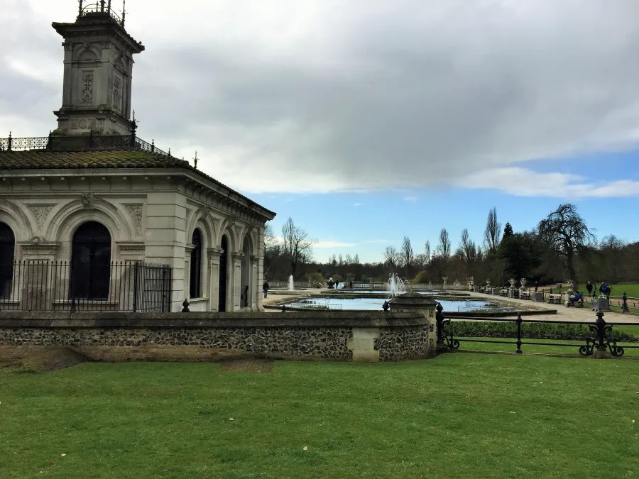 Blick auf ein historisches Gebäude mit einer Kuppel und angrenzendem Teich, umgeben von grünem Gras und Bäumen unter einem teilweise bewölkten Himmel.