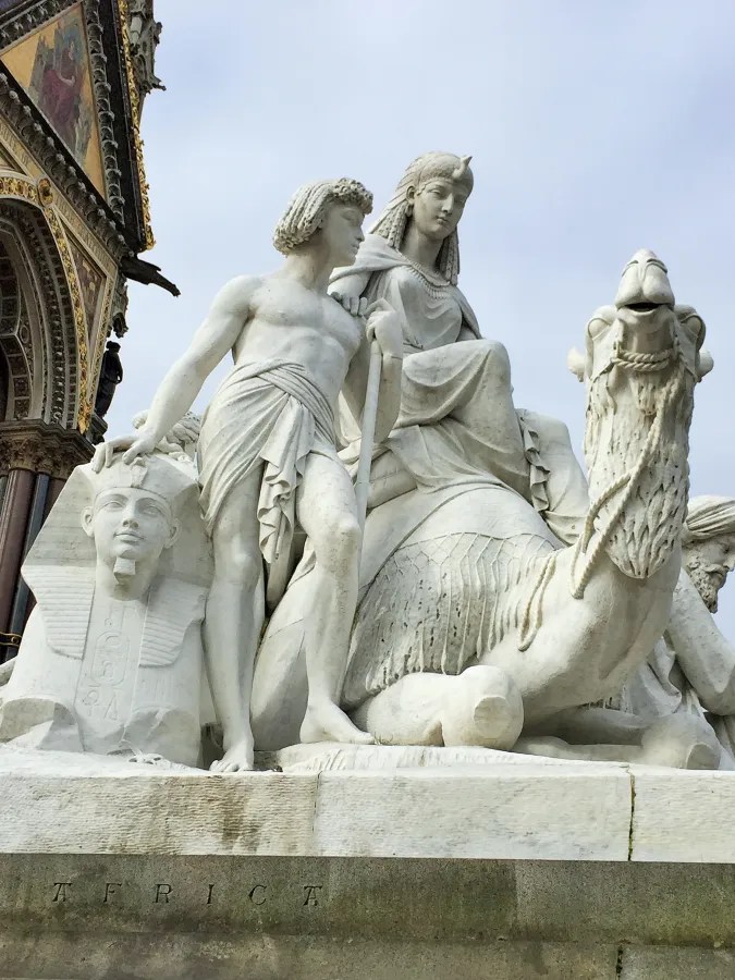 Skulptur einer weißen Steinfigur, die einen männlichen und einen weiblichen Charakter zeigt, sowie einen sitzenden Kamelkopf und eine Pharaonenstatue, vor einem historischen Gebäude.