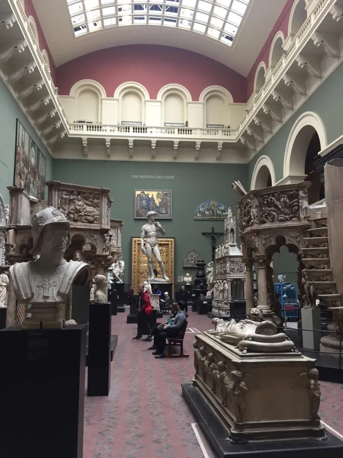 Ansicht des Innenraums des Victoria and Albert Museum, mit zahlreichen Skulpturen und Kunstwerken. Menschen sitzen und stehen zwischen den Ausstellungsstücken. Ein Highlight ist die Statue von David auf einem Podest.