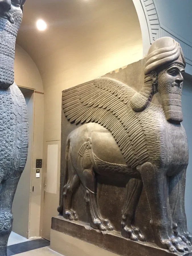 Skulptur eines geflügelten Löwen mit menschlichem Kopf, die in einem Museum ausgestellt ist. Die monumentale Statue zeigt detaillierte Texturen und Verzierungen auf dem Körper des Löwen.