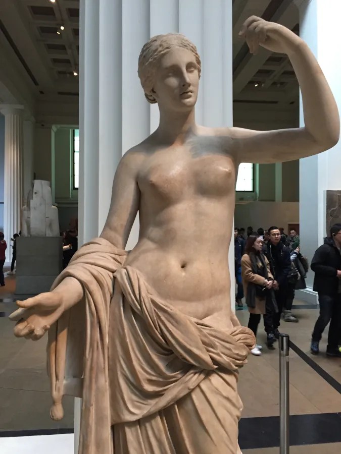 Eine antike Statue einer weiblichen Figur steht in einem Museum. Die Statue hat eine detaillierte Darstellung des Körpers und trägt einen teilweisen Gewand. Im Hintergrund sind andere Besucher und Säulen des Museums zu sehen.