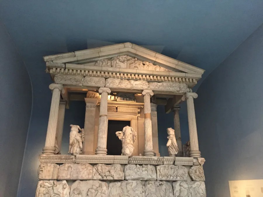 Antikes Modell eines griechischen Tempels mit Säulen und Figuren, dargestellt auf einem blauen Hintergrund.