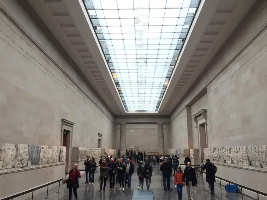 Innenansicht des British Museum in London mit Besuchern, die die Ausstellungen betrachten. An den Wänden sind antike Skulpturen sowie Reliefs zu sehen.