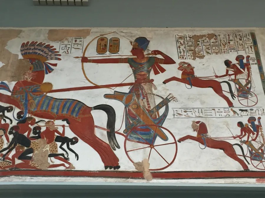 Ein altes ägyptisches Wandgemälde, das einen Krieger in einem Streitwagen zeigt, begleitet von weiteren Figuren und Tieren, mit hieroglyphischen Inschriften im Hintergrund.