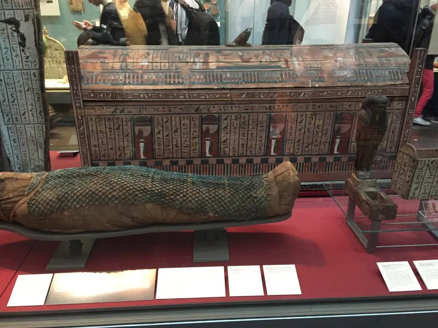 Ein antikes ägyptisches Mumien-Exponat im British Museum, umgeben von weiteren historischen Artefakten und Informationen im Hintergrund.