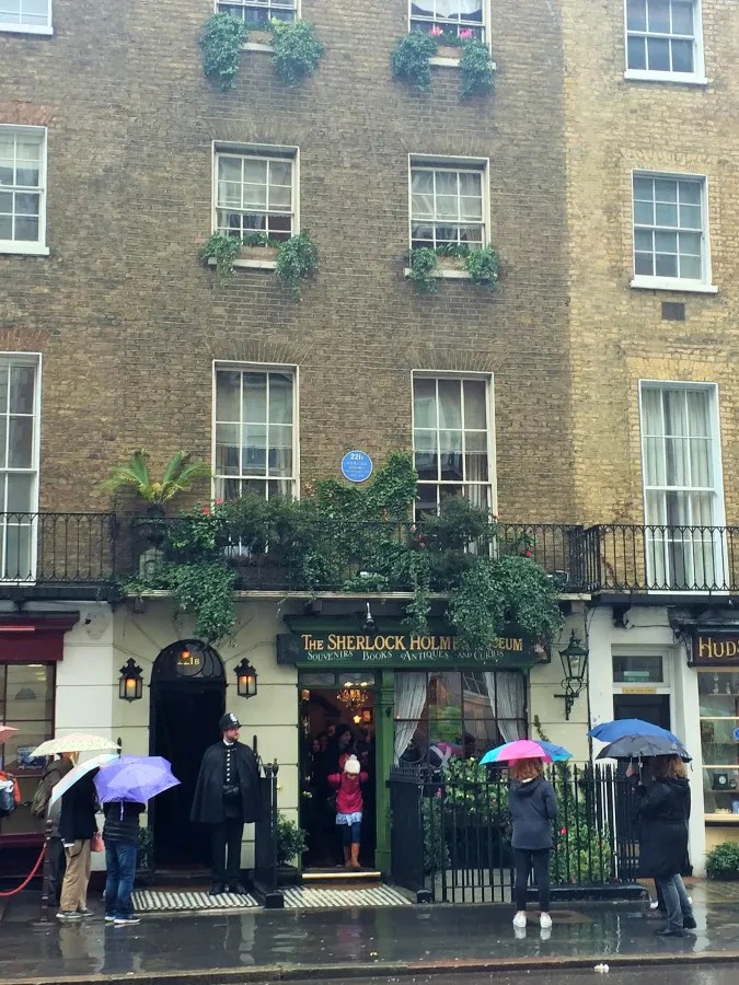 Das Sherlock Holmes Museum in London, mit Touristen, die beim Eingang Schlange stehen und Regenschirme benutzen. Die Fassade des Gebäudes ist mit Pflanzen dekoriert.
