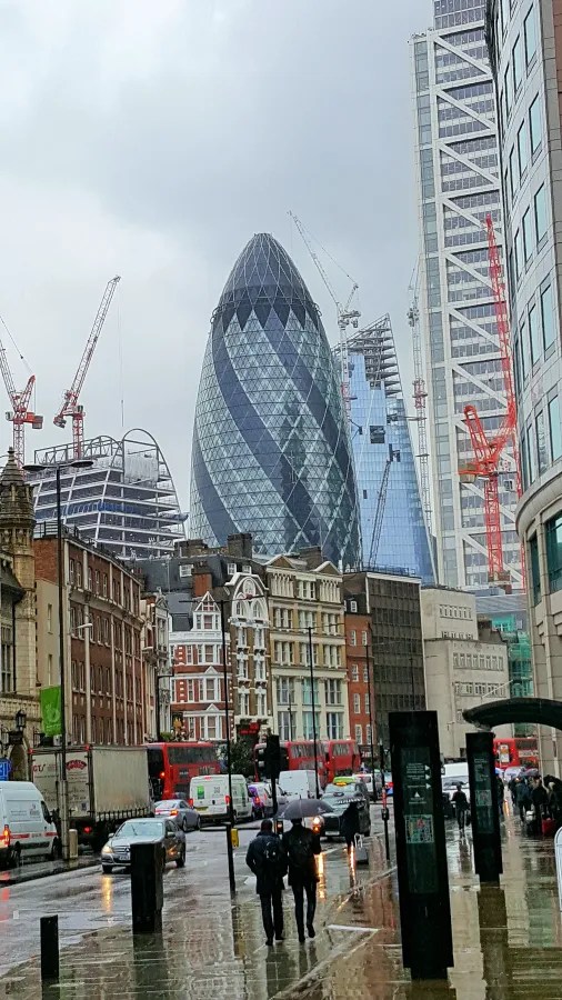 Stadtansicht von London mit modernen Gebäuden wie dem Gherkin (Schäkel) und Baumaschinen, bei grauem Himmel und Regen.