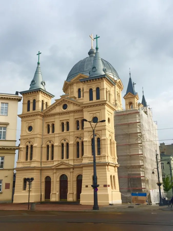 Ansicht einer historisch anmutenden Kirche in Łódź mit geschwungenen Dächern und kreuzförmigen Türmen, teilweise mit Gerüst bedeckt.
