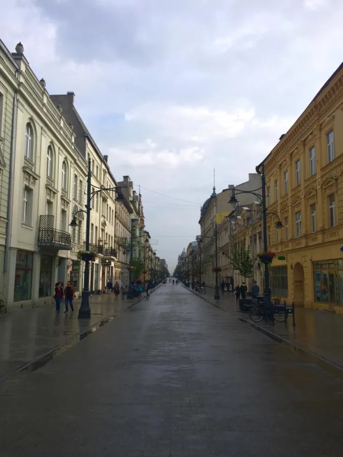 Eine belebte Straße in Łódź mit Gebäuden im historischen Stil, nassen Pflastersteinen und einem bewölkten Himmel.