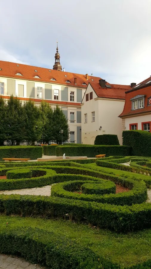 Ein grüner Park mit kunstvoll geschnittenen Hecken und einem zentralen Wasserbrunnen, umgeben von Gebäuden mit roten Dächern und Bäumen.