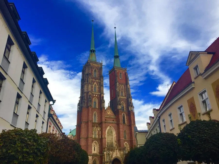 Die beeindruckende Kathedrale in Breslau mit zwei hohen Türmen und grünlichen Spitzen, umgeben von historischen Gebäuden und einem blauen Himmel.