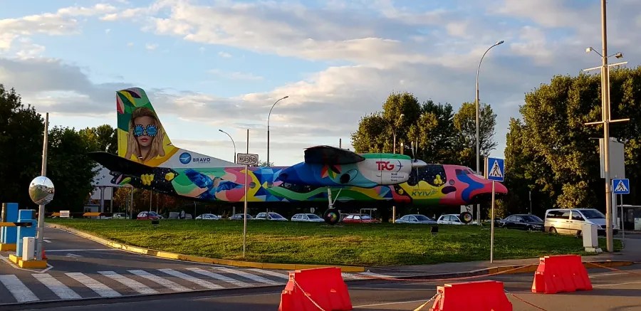 Ein bunt bemaltes Flugzeug steht auf einem Kreisverkehr, umgeben von Bäumen und Verkehrsschildern. Im Hintergrund sind Autos zu sehen und der Himmel zeigt einen dämmernden Lichtton.