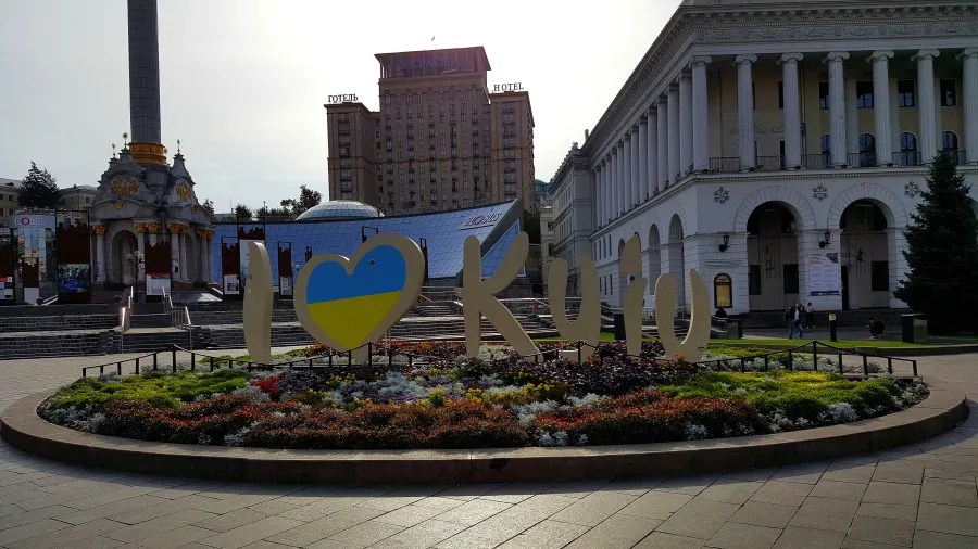Ein großer, bunten Schriftzug "I ❤️ Kyiv" auf einem Platz mit Blumenbeeten und einem historischen Gebäude im Hintergrund.