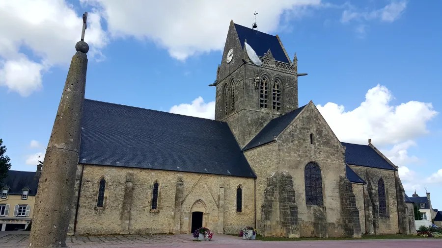Blick auf die Kirche in Sainte-Mère-Église mit einem charakteristischen Kirchturm und einem klaren blauen Himmel im Hintergrund.