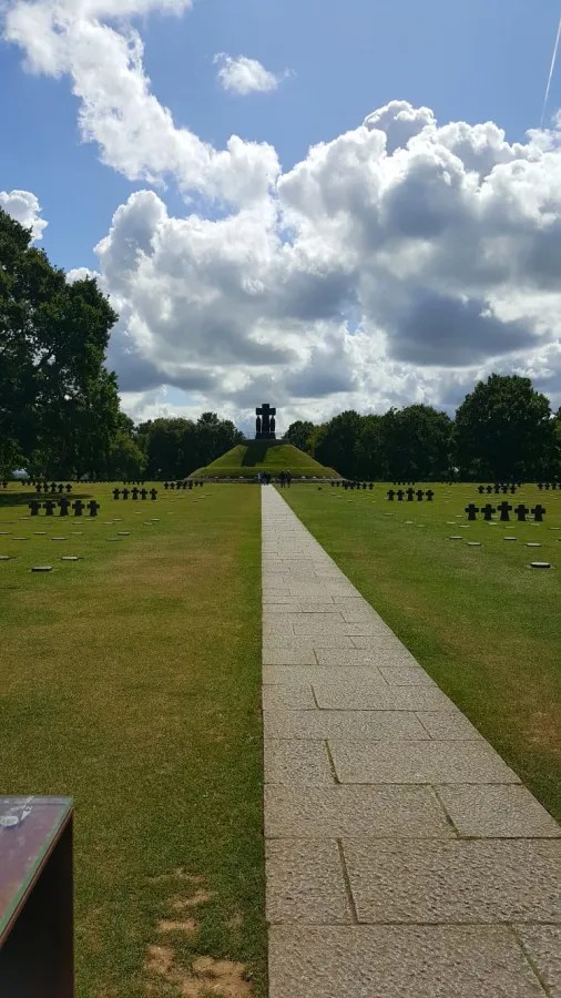 Ein Blick auf den deutschen Soldatenfriedhof in La Cambe mit dem Gedenkmonument im Hintergrund, umgeben von weißen und schwarzen Grabsteinen auf einer grünen Wiese.