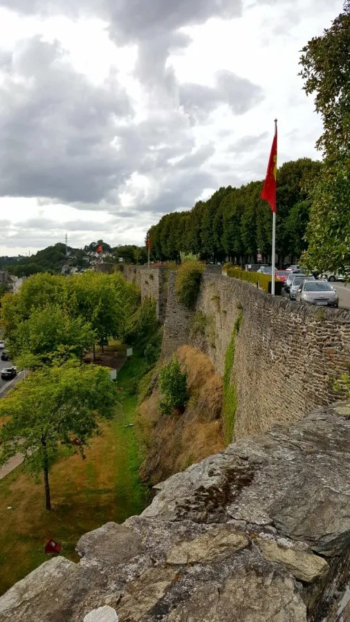Blick auf die Stadtmauer von Saint-Lô mit Bäumen und Autos im Vordergrund, bewölkter Himmel.