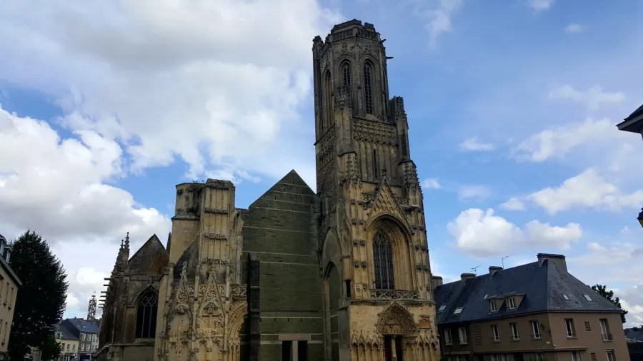 Historische Kirche in Sainte-Mère-Église mit gotischer Architektur und beeindruckendem Turm unter einem wolkigen Himmel.