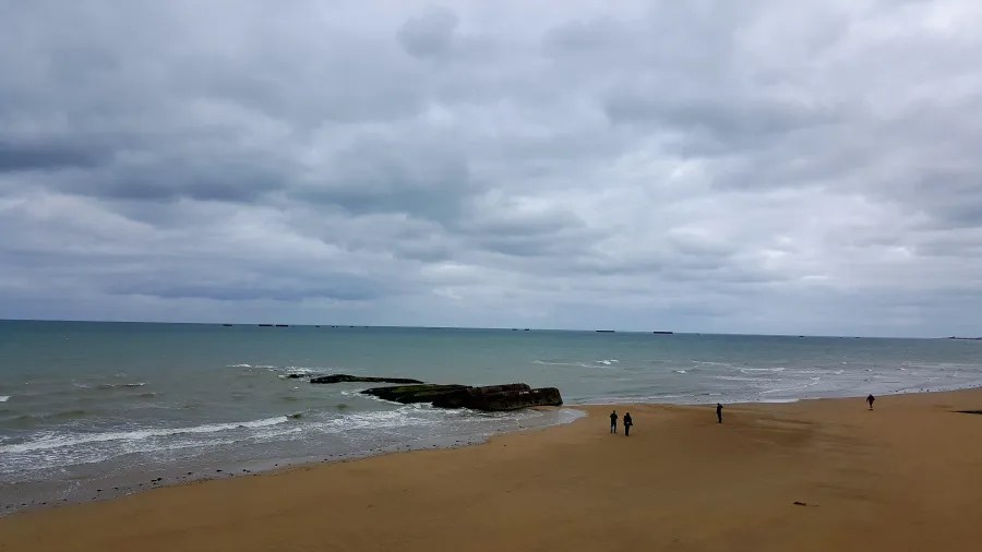 Blick auf einen Strand mit rauem Wasser und bewölktem Himmel, während zwei Personen am Ufer spazieren.