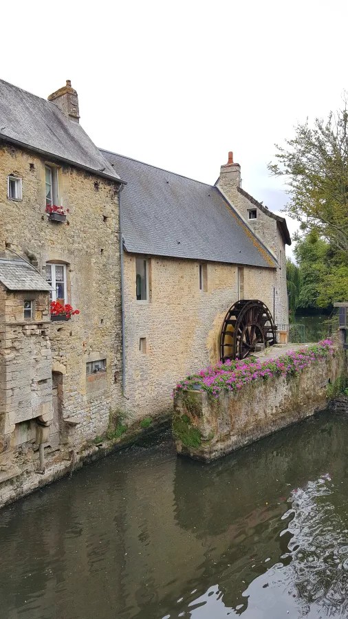 Ein historisches Gebäude mit einem Wasserrad, das an einem ruhigen Fluss in einer malerischen Umgebung steht. Bunte Blumen schmücken die Mauer des Gebäudes.