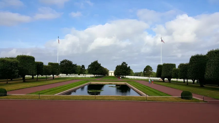 Panoramablick auf den US-Friedhof in Colleville-sur-Mer, mit frisch geschnittenen Hecken, Flaggen und einem Wasserbecken im Vordergrund.