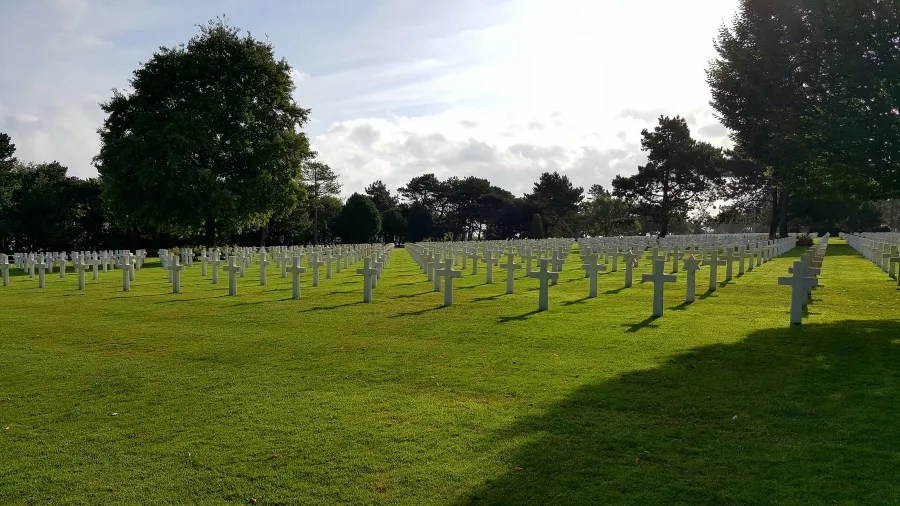 Ein Bild eines weitläufigen Friedhofs mit weißen Kreuzgräbern, umgeben von grünem Rasen und vereinzelten Bäumen, unter einem bewölkten Himmel.