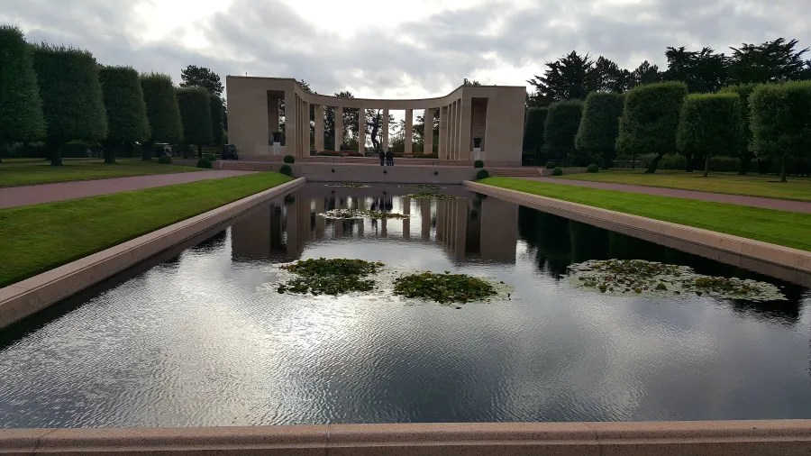 Eine friedliche Gedenkstätte mit einem zentralen Wasserbecken, umgeben von trimmed Bäumen und einem monumentalen Bauwerk im Hintergrund.